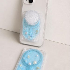 Puede incluir: Una funda transparente para smartphone con un compartimento lleno de líquido azul claro que contiene pequeños dijes de conchas blancas, diminutos detalles de palmeras doradas y cuentas iridiscentes, con un agarre de concha blanca.