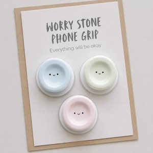 May include: Three circular phone grips in pastel blue, pale green, and soft pink, each featuring a small, simple smiling face. They are mounted on a white display card with the text WORRY STONE PHONE GRIP and Everything will be okay.
