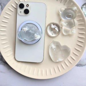 May include: A white smartphone with a circular cloud-shaped resin phone grip attached to the back. Beside the phone are three clear resin pieces: a small circular disc, a bear-shaped figure, and a faceted heart, all containing white inclusions.