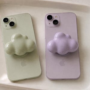 May include: Two smartphones, one light green and one light purple, each featuring a glossy, three-dimensional cloud-shaped phone grip attached to the back. The devices display the Apple logo on the rear casing and have dual camera lenses.
