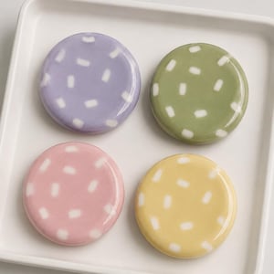 May include: Four round ceramic coasters in pastel shades: lavender, green, pink, and yellow. Each coaster has a glossy finish and is decorated with white rectangular dashes. The coasters are arranged on a white rectangular tray.