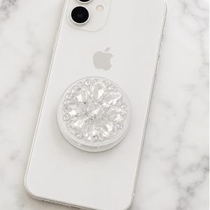 May include: A white smartphone with a circular, clear, and silver-toned phone grip attached to the back. The grip features a decorative, flower-like arrangement of faceted, clear, crystal-style stones that reflect light with a brilliant shine.