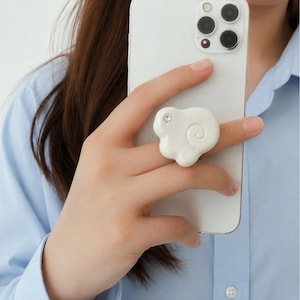 May include: A white, cloud-shaped phone grip with a glossy finish and a single embedded clear rhinestone. The accessory is attached to the back of a white smartphone, featuring a spiral design detail on its surface and a rounded, irregular shape.