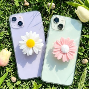 May include: Two mobile phone cases with daisy flower designs. One is lavender with a white daisy and yellow centre. The other is mint green with a pink daisy and white centre. The cases are on green grass with flowers.