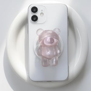 May include: A white smartphone with a clear case and a pink bear-shaped phone grip. The bear grip is translucent and has a glossy finish. The phone is resting on a white, curved surface.