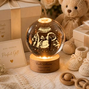 Puede incluir: Un adorno esférico de cristal con una imagen de ecografía grabada, iluminado sobre una base de madera circular con el texto grabado Noah Williams, Baby Coming, August 5, 2026. Incluye una caja de regalo, un oso de peluche y patucos.