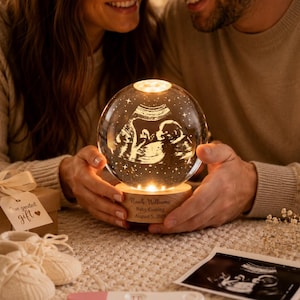 May include: A spherical glass night light featuring an etched ultrasound image, resting on a circular wooden base engraved with the text Noah Williams, Baby Coming, August 5, 2026. The light emits a warm glow, illuminating the internal design.