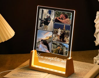 Op de afbeelding: Een houten fotolijst met een heldere acryl inzet die een collage van foto's toont. De lijst heeft een warm wit licht dat de foto's verlicht. De tekst "You complete me" is in de lijst gegraveerd.