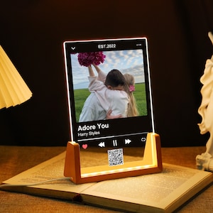 Könnte beinhalten: Ein Nachtlicht aus Holz mit einem Musikplayer, das eine klare Acrylscheibe mit einem Foto eines Paares und dem Songtitel "Adore You" von Harry Styles zeigt. Der Player hat ein Herzsymbol, Play-, Pause- und Vorwärtstasten sowie einen QR-Code.