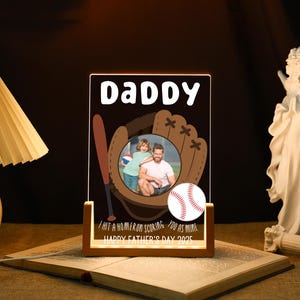 Könnte beinhalten: Ein beleuchteter Acryl-Fotorahmen mit Baseball-Motiv. Der Rahmen zeigt das Wort "Daddy" oben, einen Baseballhandschuh, einen Baseballschläger und einen Baseball. Der Rahmen enthält ein Foto eines Mannes und eines Kindes. Der Text unten lautet "I hit a homerun scoring you as mine. Happy Father's Day 2025."