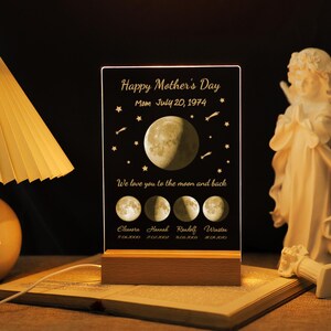 Könnte beinhalten: Ein personalisiertes Nachtlicht zum Muttertag mit Mond- und Sternenmotiv. Die schwarze Acrylplatte trägt den Text "Happy Mother's Day" und "We love you to the moon and back". Es enthält einen Holzsockel und eine Vollmondgrafik.