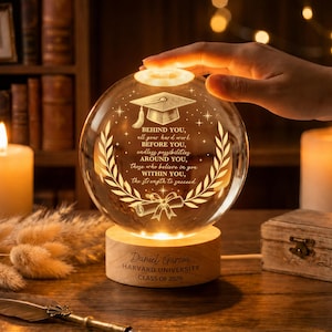 Op de afbeelding: Een heldere glazen afstudeerbol met een houten voet, gegraveerd met een boodschap en de tekst "Daniel Garcia, Harvard University, Klasse 2026." De bol is van binnenuit verlicht en een hand raakt de bovenkant aan. De bol heeft een ontwerp met afstudeerhoed en lauwerkrans.