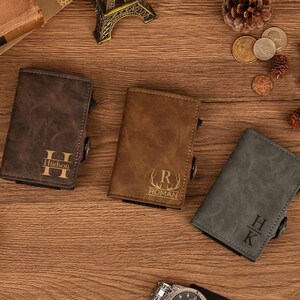 Op de afbeelding: Drie gepersonaliseerde leren portemonnees in bruin, beige en grijs, elk met een unieke initialen- en naamgravering. De portemonnees worden gepresenteerd op een houten oppervlak, waarbij hun textuur wordt getoond.