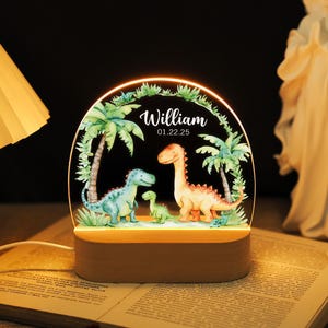 Peut inclure: Veilleuse personnalisée avec une famille de dinosaures bleus, verts et oranges, sur fond de palmiers. Le nom "William" et la date "01.22.25" sont affichés sur la lumière. La base est en bois.