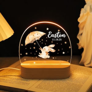 May include: A personalised night light with a watercolour illustration of a bunny holding an umbrella, with the name "Easton" and date "11.18.25" printed on the acrylic. The base is made of wood, and the light emits a warm glow.
