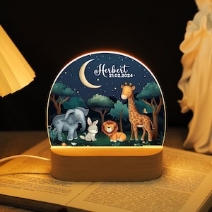May include: A wooden night light with a round, illuminated panel featuring a moon, stars, and cartoon animals including an elephant, rabbit, lion, and giraffe. The panel also includes the name "Herbert" and the date "21.02.2024".