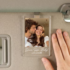May include: A clear acrylic car visor photo holder featuring a photograph of a couple. The text "Drive Safe, Baby!" and "LOVE, ANNE" is printed on the bottom. The holder is attached to a car sun visor.