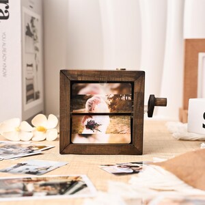 Könnte beinhalten: Ein hölzerner Fotowürfel in Dunkelbraun mit Drehmechanismus, der ein Hochzeitsfoto zeigt. Der Würfel hat einen kleinen Holzgriff. Mehrere gedruckte Fotos liegen auf einer hellen Oberfläche um den Würfel herum.