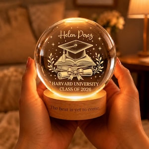Op de afbeelding: Een heldere glazen sneeuwbol met een houten voet, van binnenuit verlicht. De bol heeft een gegraveerd ontwerp van een afstudeerhoed, boeken en een rol, met de tekst "Helen Perez" en "Harvard University Class of 2026." De voet luidt "The best is yet to come."