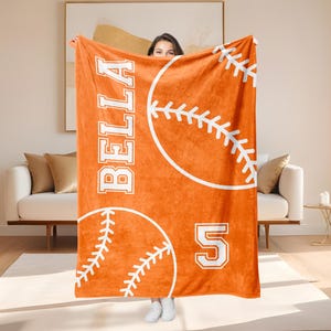 May include: An orange blanket featuring a baseball design with white stitching and the name "BELLA". The number "5" is also displayed. The blanket is held up by a person.
