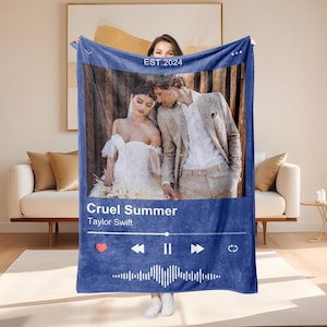 Pode incluir: Um cobertor azul marinho com um design que imita uma interface de reprodutor de música, exibindo uma foto de um casal. O cobertor inclui o título da música "Cruel Summer" de Taylor Swift, junto com um botão de reproduzir/pausar e uma forma de onda. O texto "EST.2024" também está presente.