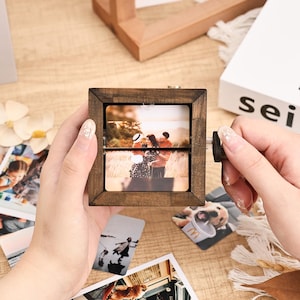 May include: A wooden photo cube with a rotating mechanism, displaying two images. The cube is held by a person, with other photos scattered around. The cube has a dark brown finish, and the photos show a couple and other memories.