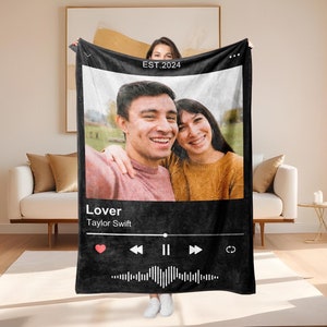 Pode incluir: Cobertor de lã preto e branco com uma foto de um casal em uma moldura preta. O cobertor tem um design de reprodutor de música com o texto "Lover" e "Taylor Swift" e o ano "EST. 2024".