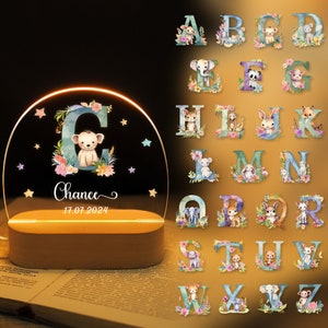 Op de afbeelding: Een nachtlampje met een heldere acrylbasis en een koepelvormige kap. De kap heeft een aquarel-illustratie van de letter C met een schattige aap en bloemen. De naam "Chance" en de datum "17.07.2024" staan onder de letter. De afbeelding toont ook een verzameling van 26 letters van het alfabet, elk met een ander dier- en bloemendesign.