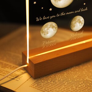 Pode incluir: Luz noturna personalizada com base de madeira e painel acr&iacute;lico transparente. O painel &eacute; gravado com ilustra&ccedil;&otilde;es de luas, nomes, datas e a frase "We love you to the moon and back."