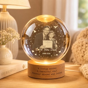 Peut inclure: Une veilleuse sphérique en verre avec une gravure laser représentant un homme et un enfant tenant un certificat. Le texte indique "Love You Dad 1968-2026" avec des étoiles et des colombes. La base en bois porte l'inscription "A loving father A beautiful soul Forever missed and loved."