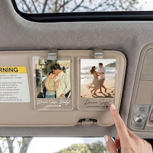 May include: Two rectangular clear acrylic photo clips attached to a car sun visor. The left clip shows a couple embracing with the text Drive Safe Baby! LOVE-ANNE. The right clip shows a couple on a beach with the text Drive Safe! ALWAYS WITH YOU.
