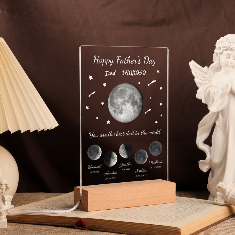Pode incluir: Um presente personalizado para o Dia dos Pais com um painel de acr&iacute;lico transparente com um design de fases da lua e o texto "Happy Father's Day Dad 17/11/1959". O painel est&aacute; sobre uma base de madeira com um cabo de alimenta&ccedil;&atilde;o. O texto "You are the best dad in the world".