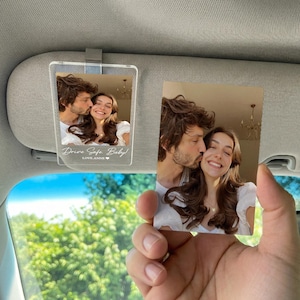 May include: A rectangular clear acrylic car visor clip featuring a printed photograph of a couple, with the white text "Drive Safe, Baby!" and "LOVE, ANNE" followed by a small heart icon printed at the bottom of the acrylic surface.