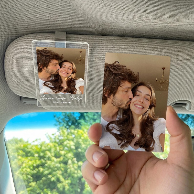 May include: A rectangular clear acrylic car visor clip featuring a printed photograph of a couple, with the white text "Drive Safe, Baby!" and "LOVE, ANNE" followed by a small heart icon printed at the bottom of the acrylic surface.
