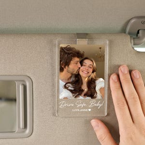 May include: A clear rectangular acrylic car visor clip featuring a printed photograph of a couple. The text "Drive Safe, Baby!" and "LOVE, ANNE" with a small heart icon are printed in white at the bottom of the transparent acrylic plaque.