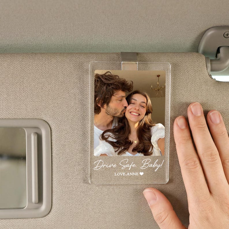 May include: A clear rectangular acrylic car visor clip featuring a printed photograph of a couple. The text "Drive Safe, Baby!" and "LOVE, ANNE" with a small heart icon are printed in white at the bottom of the transparent acrylic plaque.