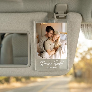 May include: A clear rectangular acrylic car visor clip featuring a printed photograph of a couple embracing. The bottom of the acrylic plaque displays the white text Drive Safe! I LOVE YOU. The clip is attached to a beige fabric car sun visor.