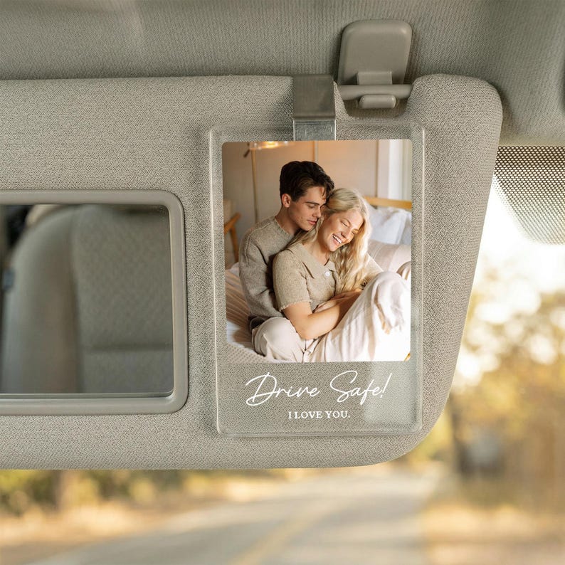 May include: A clear rectangular acrylic car visor clip featuring a printed photograph of a couple embracing. The bottom of the acrylic plaque displays the white text Drive Safe! I LOVE YOU. The clip is attached to a beige fabric car sun visor.