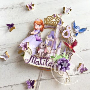 Può includere: Topper per torta viola e oro con un personaggio di una principessa, un castello, un coniglio, farfalle e il nome "Matilda" con il numero "8" su uno stendardo.