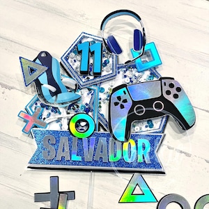 Op de afbeelding: Een blauw en zilveren taarttopper met een gamingthema. Het heeft een controller, koptelefoon en het nummer 11. De naam "SALVADOR" staat op een banner. Verschillende game-symbolen en -vormen zijn ook inbegrepen.
