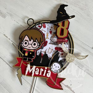 Könnte beinhalten: Ein Harry-Potter-Tortenaufsatz mit einem Cartoon-Bild von Harry Potter, einer goldenen Zahl 8, einem schwarzen Hexenhut und einer weißen Eule. Der Aufsatz enthält auch ein rotes Band mit dem Namen "Maria" in goldenen Buchstaben.