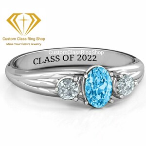 May include: A silver ring with three blue gemstones. The center stone is oval shaped and the two side stones are round. The ring is engraved with the text "CustomClassRingShop CLASS OF 2022".