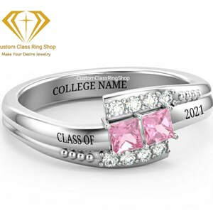 May include: A silver class ring featuring two pink square gemstones and a row of smaller clear gemstones. The ring is engraved with "COLLEGE NAME", "CLASS OF", and "2021".
