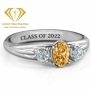 May include: A silver ring with three stones. The center stone is a large oval-shaped topaz. The two side stones are smaller round-shaped diamonds. The ring is engraved with the text "CustomClassRingShop CLASS OF 2022".