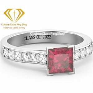 May include: A silver ring with a red square gemstone and a row of small clear gemstones around the band. The inside of the band is engraved with "CustomClassRingShop CLASS OF 2022".