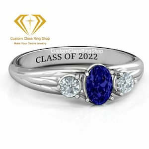 May include: A silver ring with three stones. The center stone is a large oval blue sapphire. The two side stones are smaller round clear stones. The ring is engraved with the text "CustomClassRingShop CLASS OF 2022".