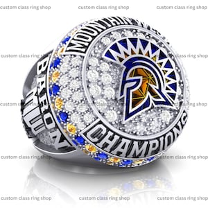 Könnte beinhalten: Silberner Ring mit detailliertem Design. Der Ring hat ein zentrales Emblem mit einem blauen und goldenen spartanischen Helm, umgeben von weißen Edelsteinen und dem Wort "CHAMPIONS". Der Ring hat auch die Wörter "MOUNTAIN" und "BARONS".