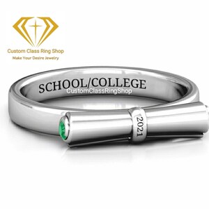 May include: A silver graduation ring with a green emerald stone and a scroll design. The ring is engraved with the text "SCHOOL/COLLEGE CustomClassRingShop 2021".
