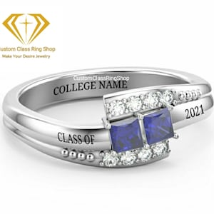 May include: A silver class ring featuring two blue gemstones and a row of smaller clear gemstones. The ring is engraved with "COLLEGE NAME", "CLASS OF", and "2021".