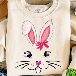 May include: A cream colored sweatshirt with a cartoon bunny face printed on the front. The bunny has pink ears, a pink bow, and black whiskers.